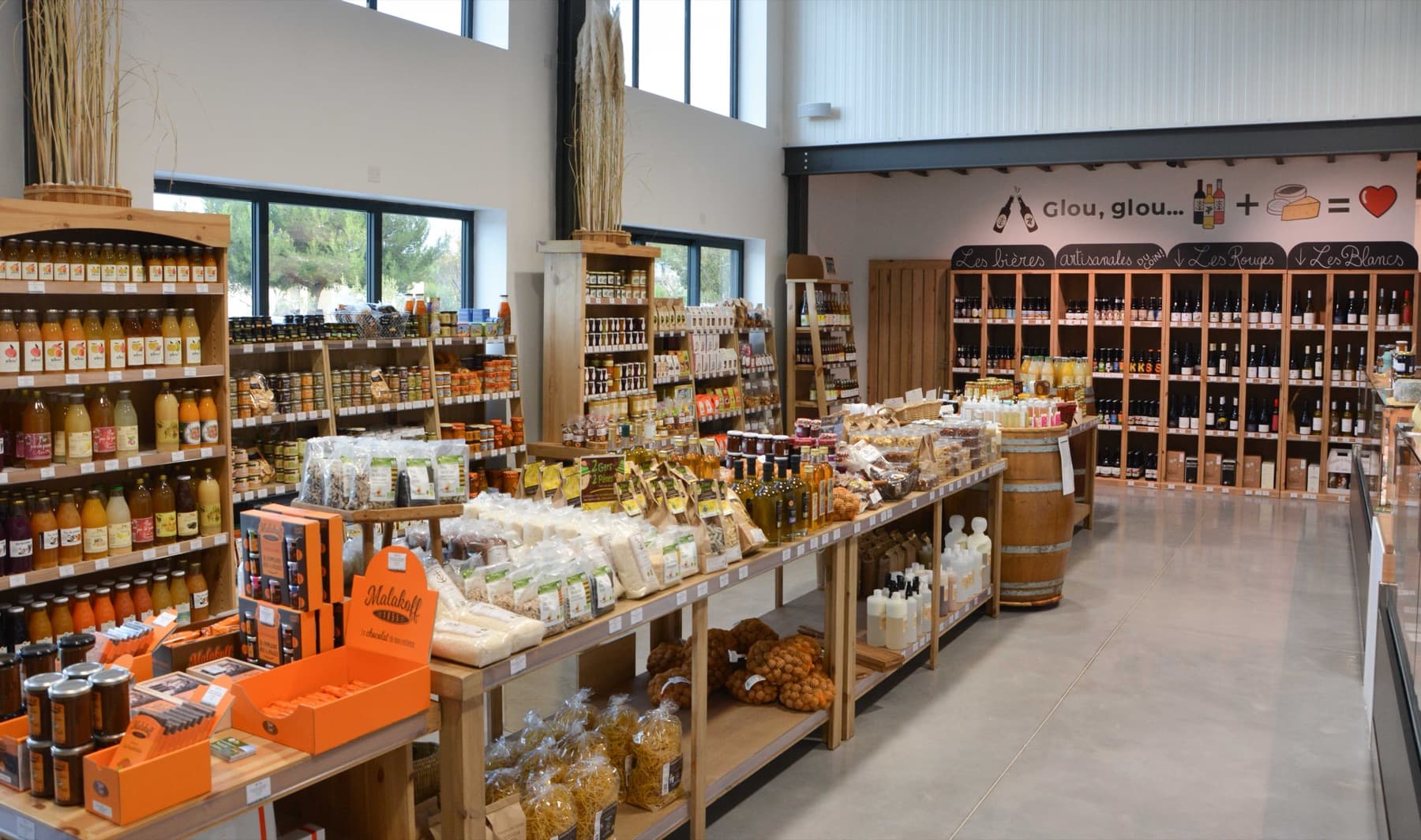 Vue large de l'épicerie du Mas du Trident à Vauvert : tables centrales de vrac, pâtes et chocolats, rayons de jus et conserves, et au fond le mur de vins et bières artisanales