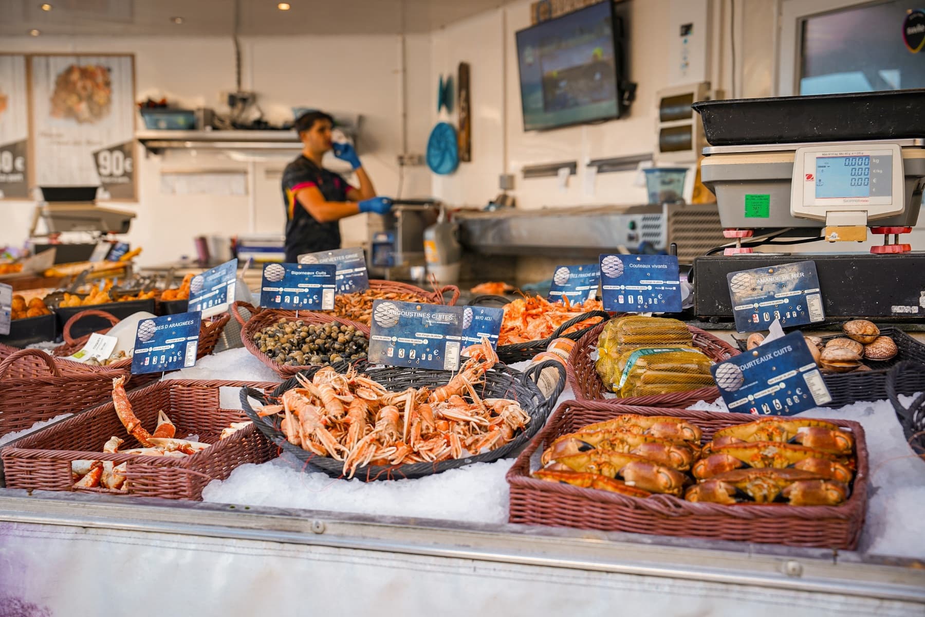 Étal réfrigéré d'une poissonnerie garni de langoustines, tourteaux, bigorneaux et coquillages sur glace pilée, chacun avec son écriteau de prix, un poissonnier en arrière-plan
